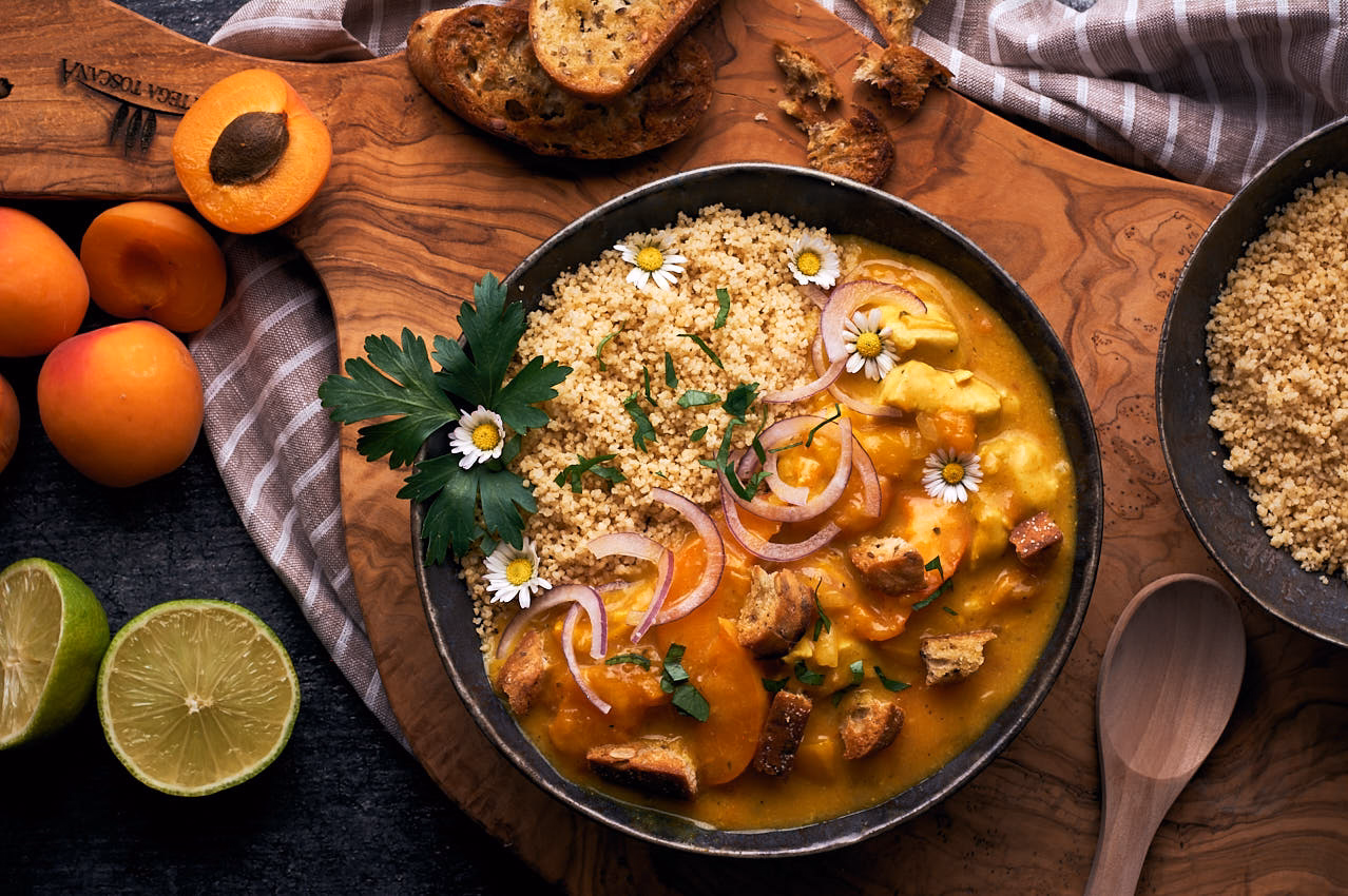 Hähnchen-Curry mit Aprikosen und Couscous, fotografiert von oben, mit Brot und Limetten im Hintergrund.
